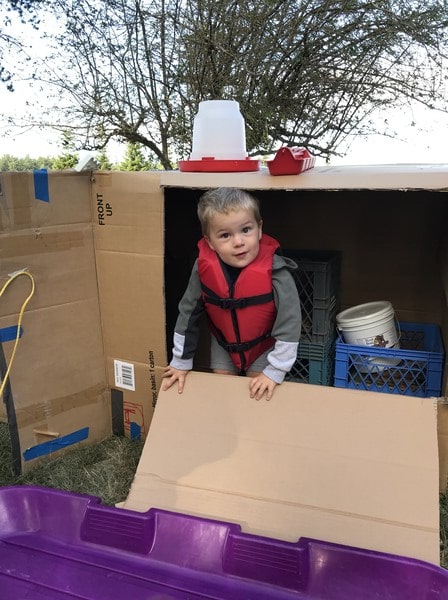 A young boy wearing a life jacket sits inside a cardboard box, ready for an adventure on the water.
